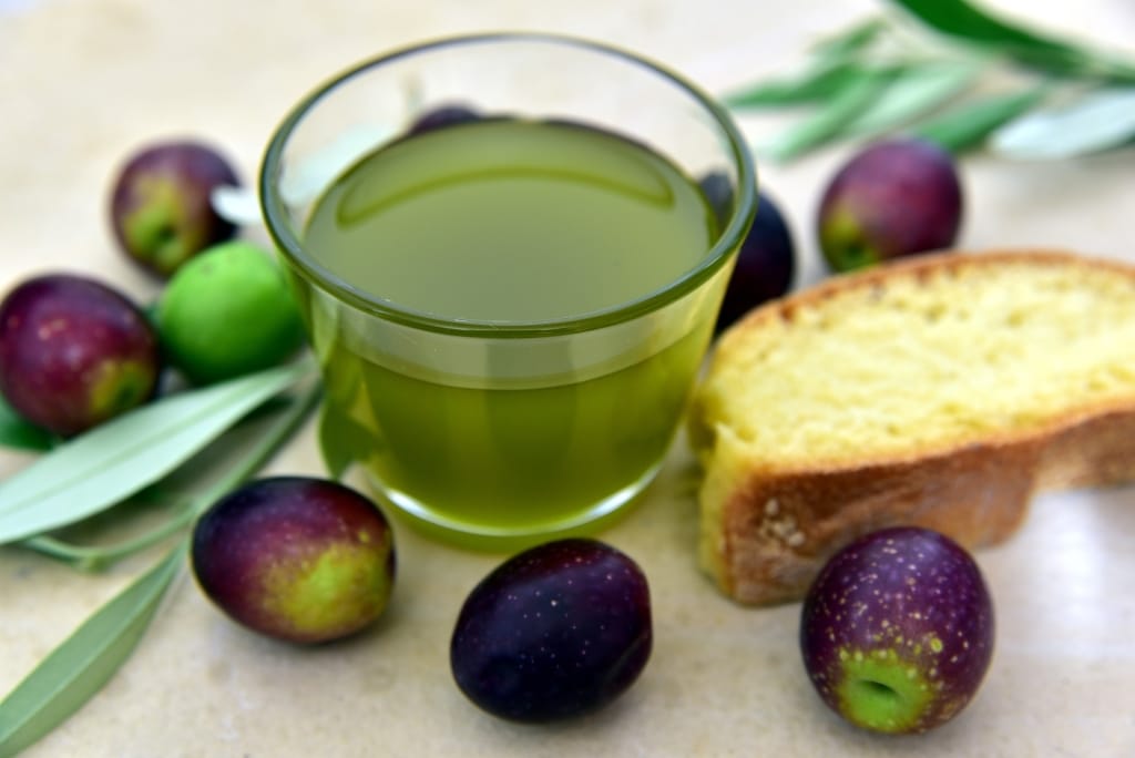 Glasschälchen mit hellgrünem Olivenöl neben grün-schwarzen Oliven und einer Scheibe Baguette
