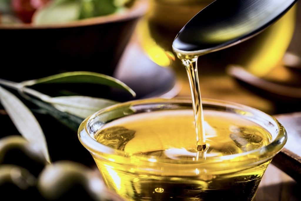 Goldenes Olivenöl fließt von einem Esslöffel in ein kleines Glasschälchen