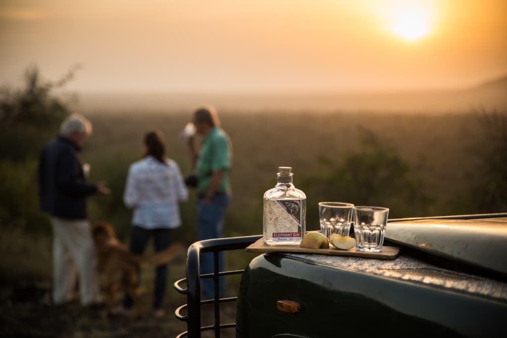 Elephant Gin mit Apfel und Gläsern auf einer Ranger-Autohaube vor Sonnenuntergang in afrikanischer Landschaft mit Menschen im Hintergrund