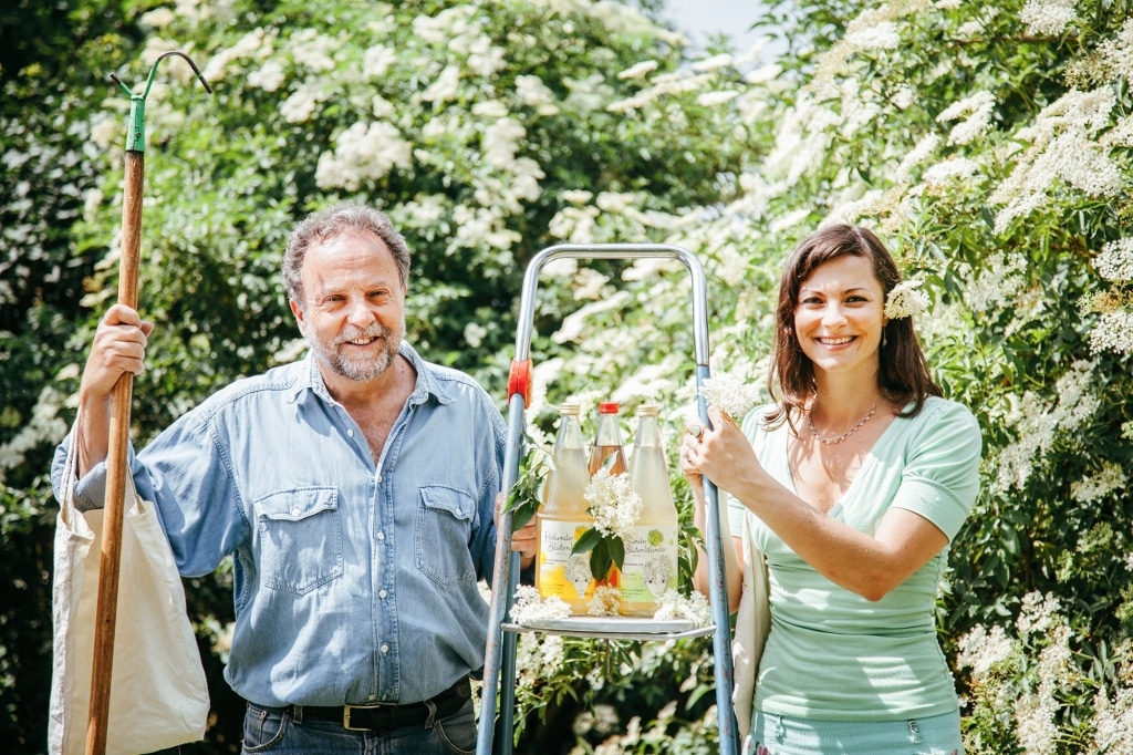 Roberto Deimel und Malena Medam von HolunderBlütenWunder stehen vor blühenden Holunderbüschen und halten eine Trittleiter mit Holunderblütensirup-Flaschen