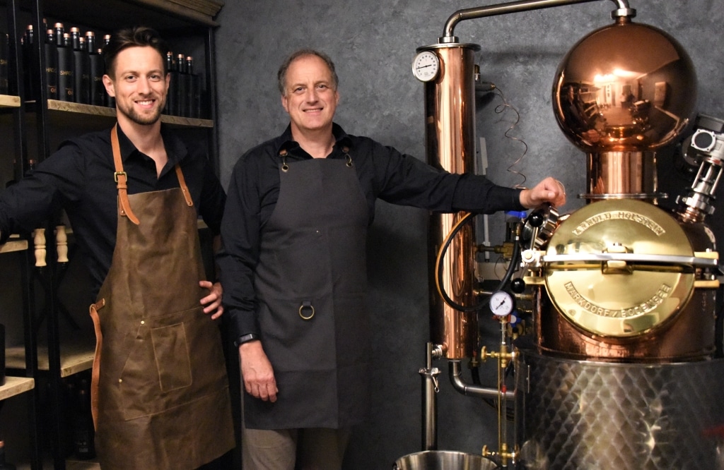 Patrick Luebke und sein Vater zwischen der Arnold Holstein Brennblase und einem Regal mit Shadows Franconian Dry Gin - Luebke & Sohn Distillers